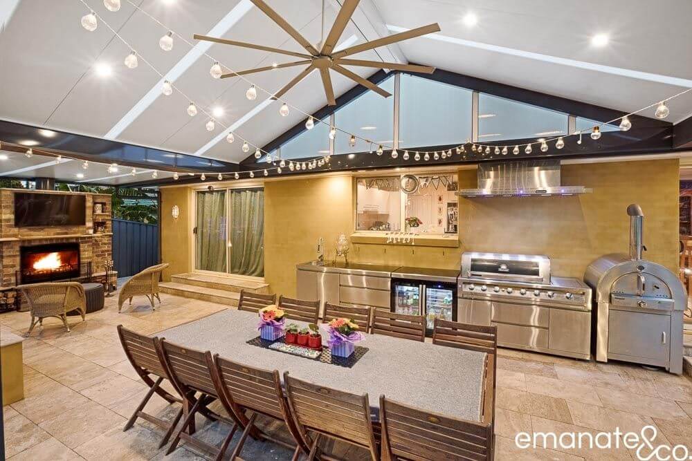 Spacious outdoor dining and cooking area with string lights, a large dining table, built-in barbecue, pizza oven, and fireplace, under a vaulted roof for year-round use.