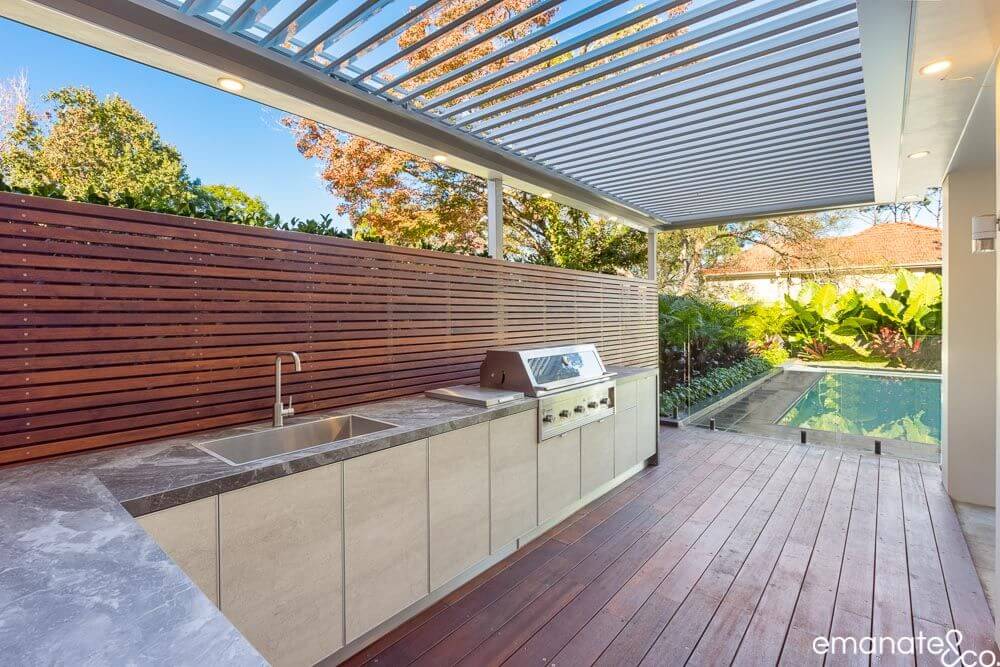 Modern outdoor kitchen with marble countertops, built-in grill, and wood privacy screen, next to a wooden deck and pool, ideal for outdoor cooking and relaxation.