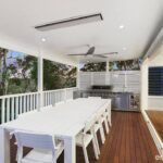 Covered outdoor dining area with a long white table and built-in barbecue, seamlessly integrated with the home's indoor kitchen and surrounded by natural greenery.