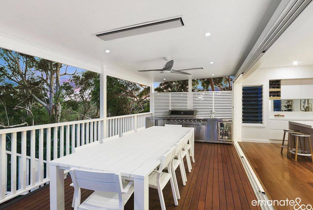 Covered outdoor dining area with a long white table and built-in barbecue, seamlessly integrated with the home's indoor kitchen and surrounded by natural greenery.