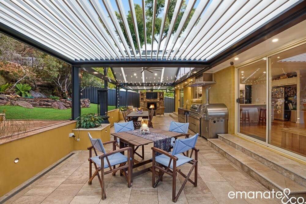 Outdoor dining area with a louvered roof and modern outdoor kitchen, featuring string lights, a built-in grill, and a cozy fireplace, perfect for entertaining.