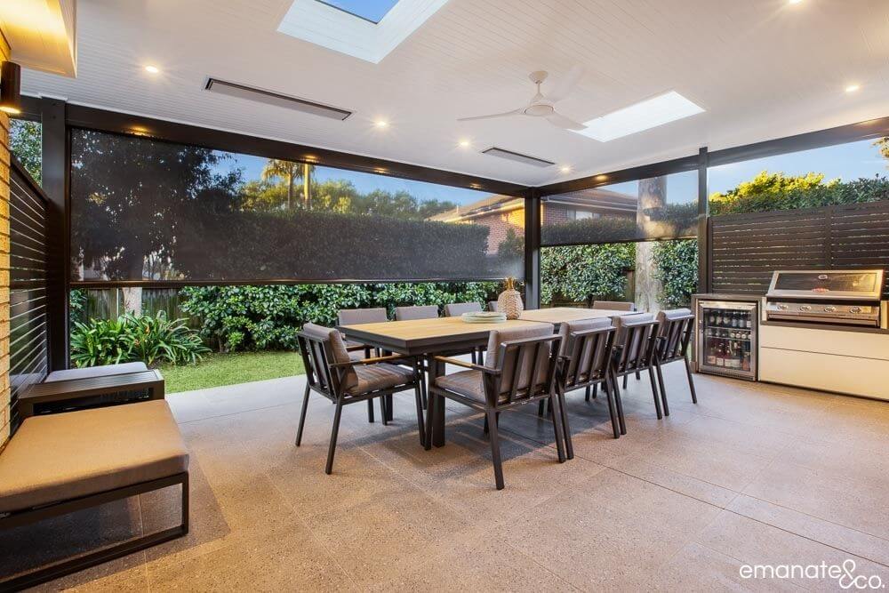 Enclosed outdoor dining area with a long table, skylights, retractable blinds, and a built-in barbecue and mini-fridge, perfect for year-round outdoor gatherings.